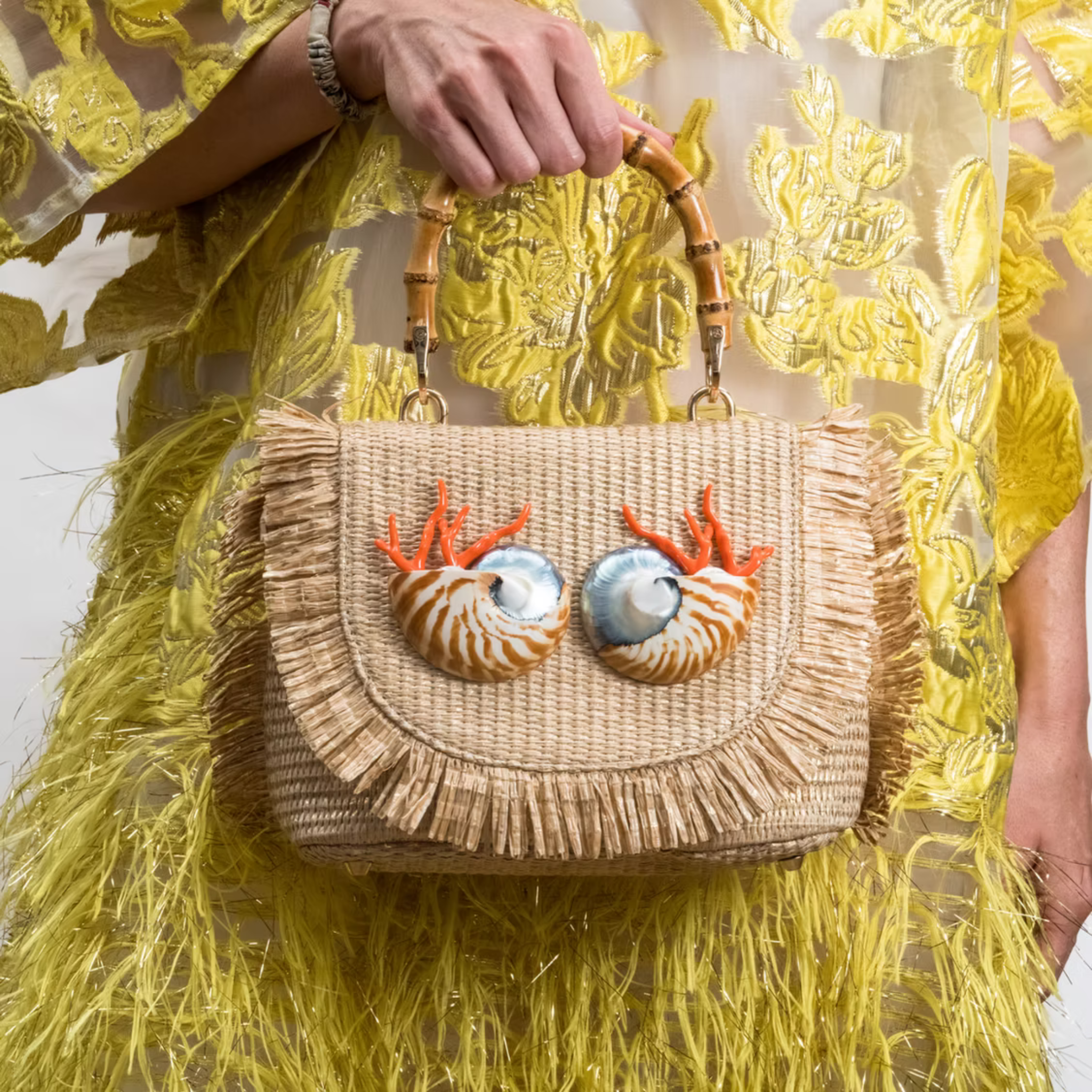 A woven straw handbag with a bamboo top handle, held by a model wearing a yellow embroidered dress. The bag features two large seashell decorations on the front flap with orange coral-like accents.