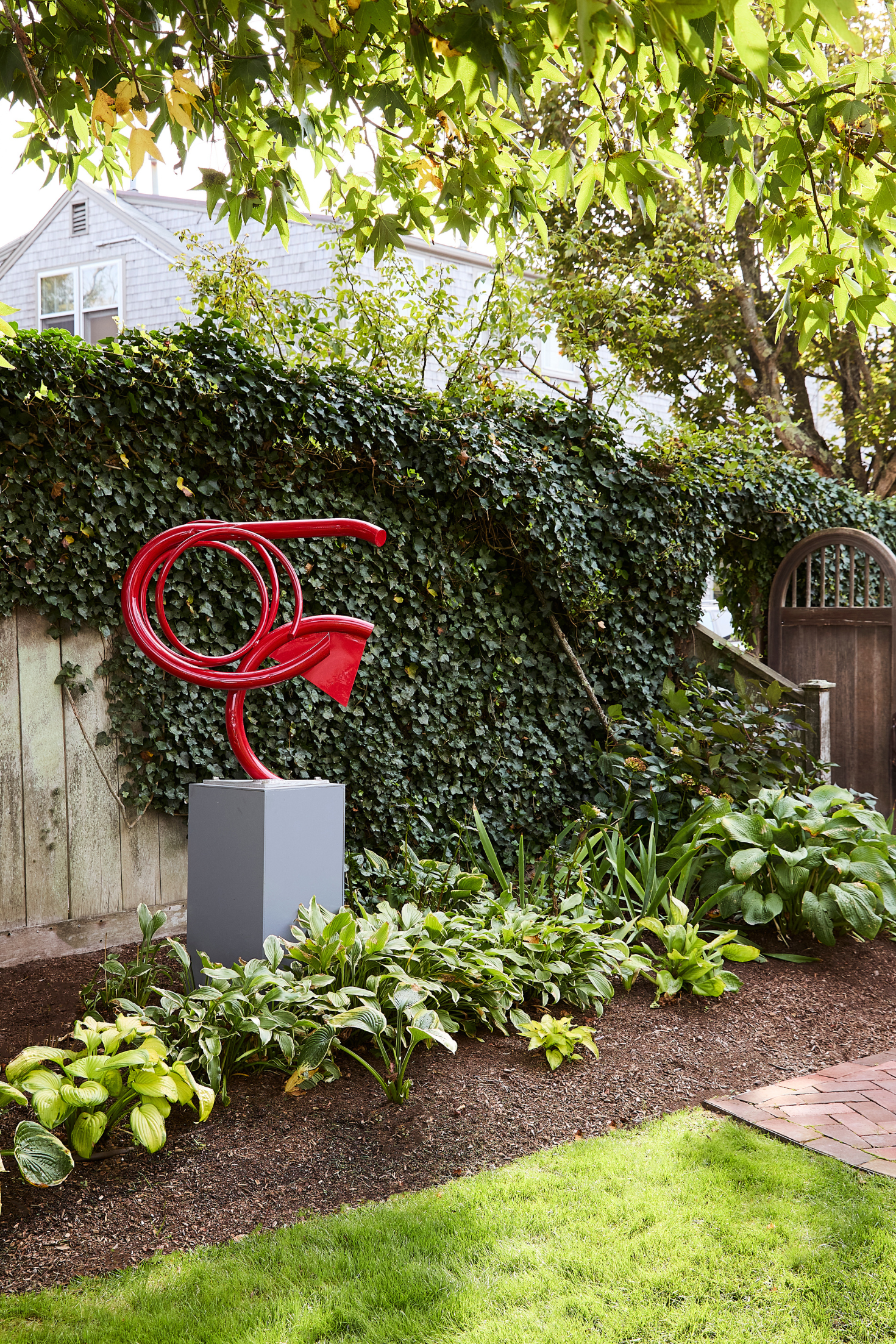 Red metal sculpture in a garden setting with greenery and a house in the background