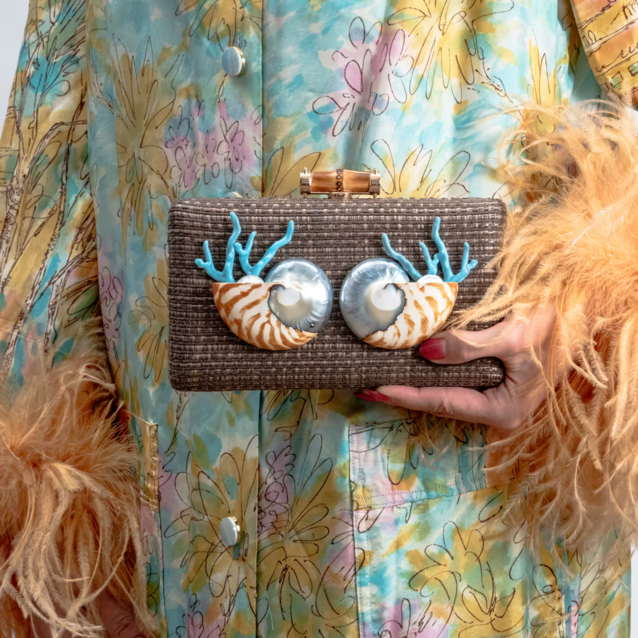 A person in a colorful floral jacket holds a brown woven clutch bag with two shell eyes and blue antler embellishments.