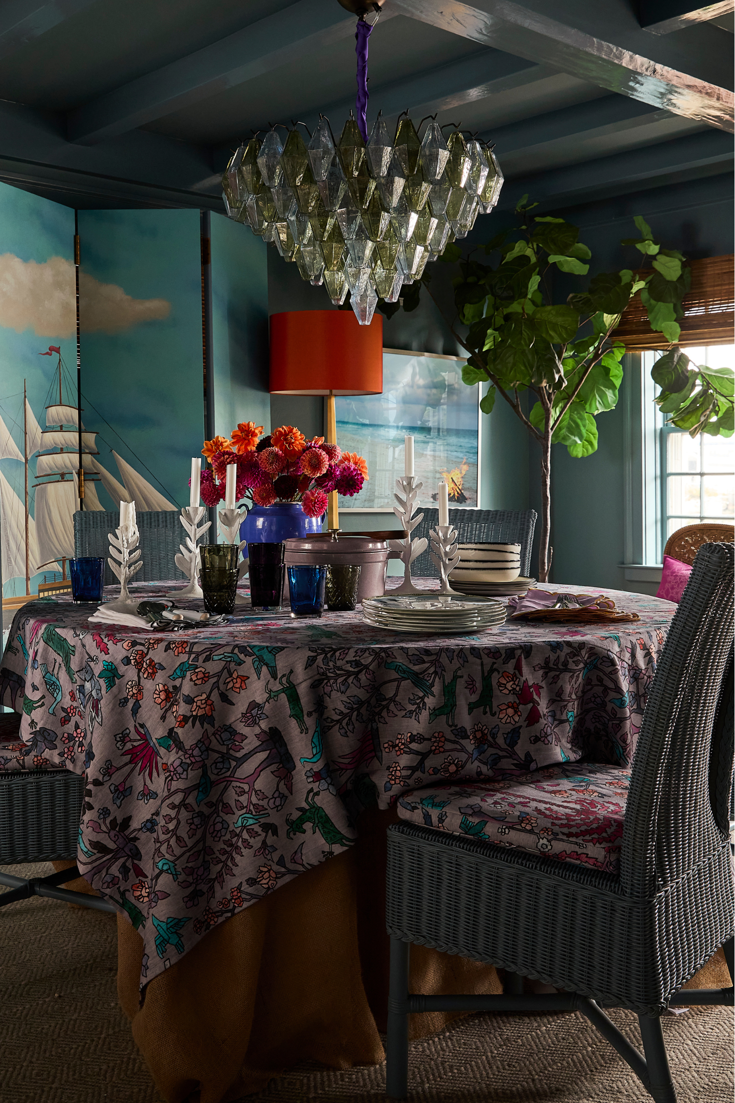 Colorful dining room with a patterned tablecloth, set table, and woven chairs beneath a geometric glass chandelier, styled with florals, candles, and coastal-inspired decor.