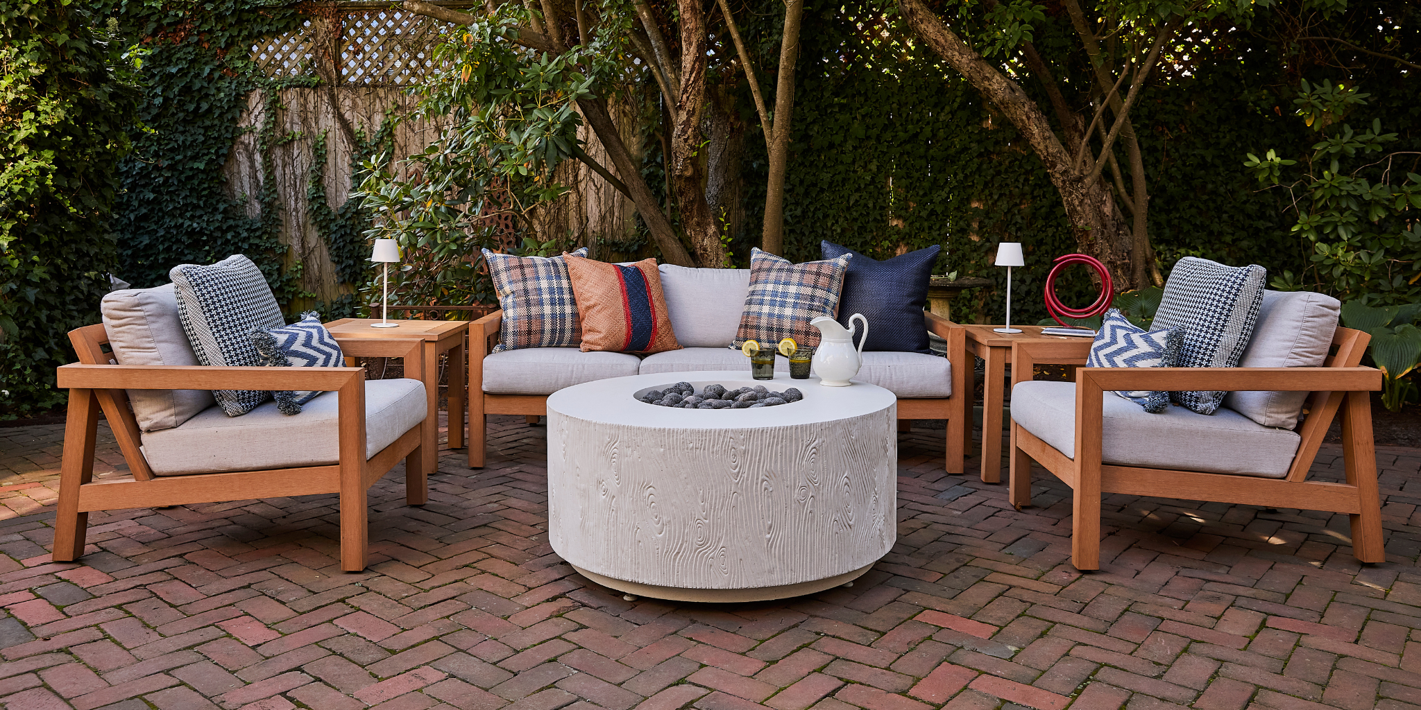 Outdoor patio set with wooden chairs, a fire pit table, and decorative pillows on a brick patio.